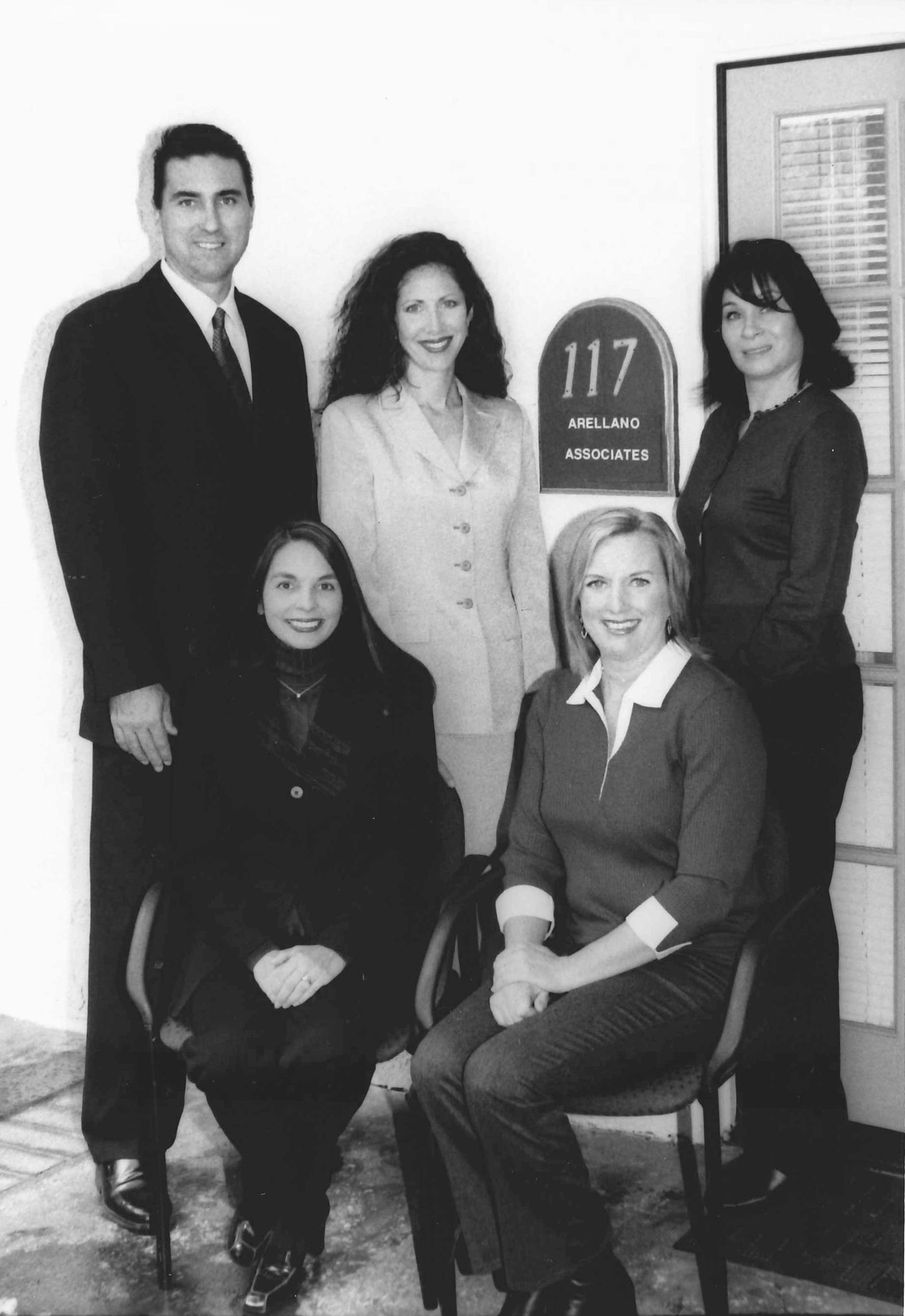 An archival black-and-white photograph of the Arellano Associates team gathered beside an office sign.