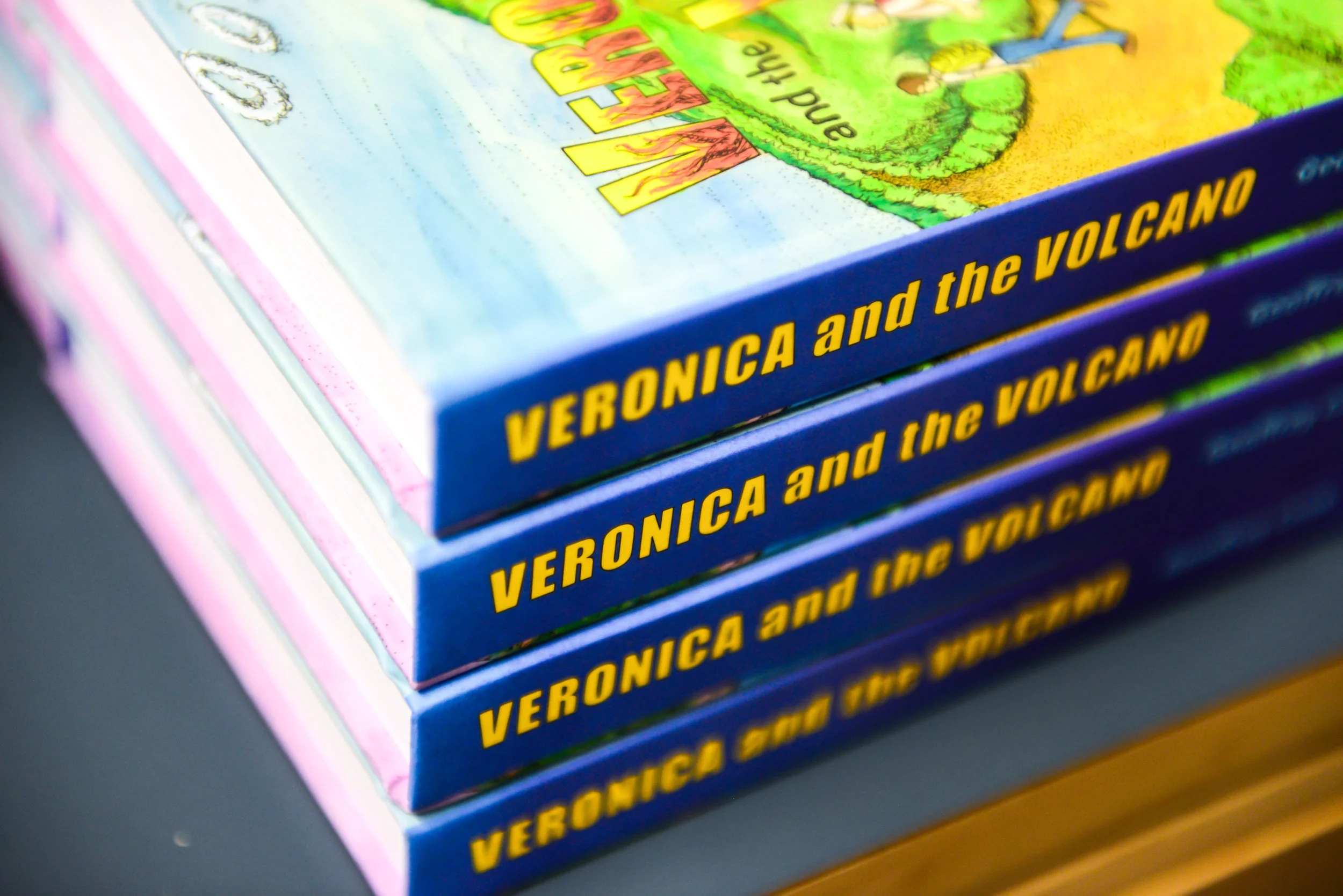 A stack of brightly illustrated copies of Veronica and the Volcano, with the title visible on their blue spines.