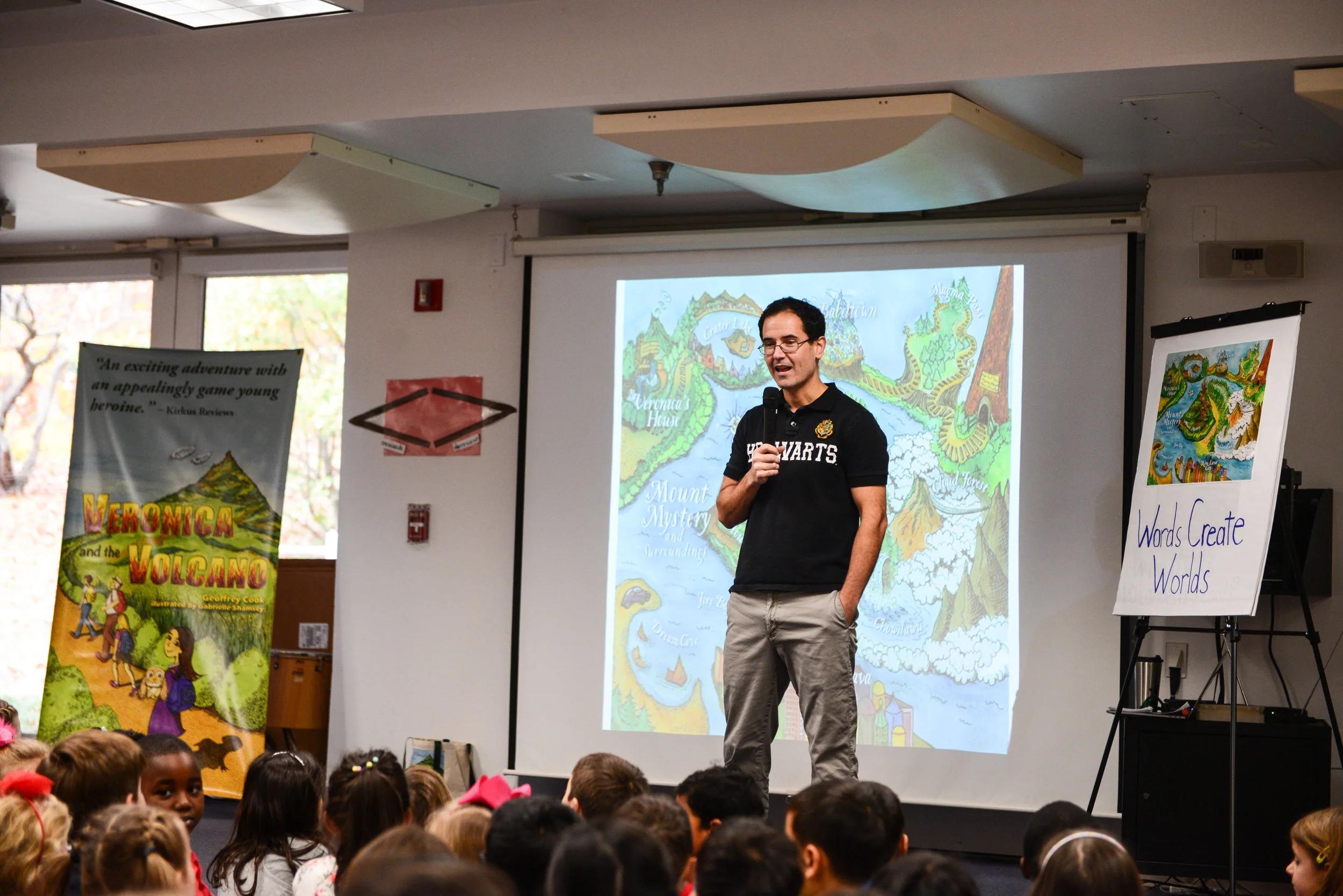 Geoffrey Cook in a Hogwarts shirt holding a microphone before an audience of schoolchildren, with a map of Veronica’s world behind him.