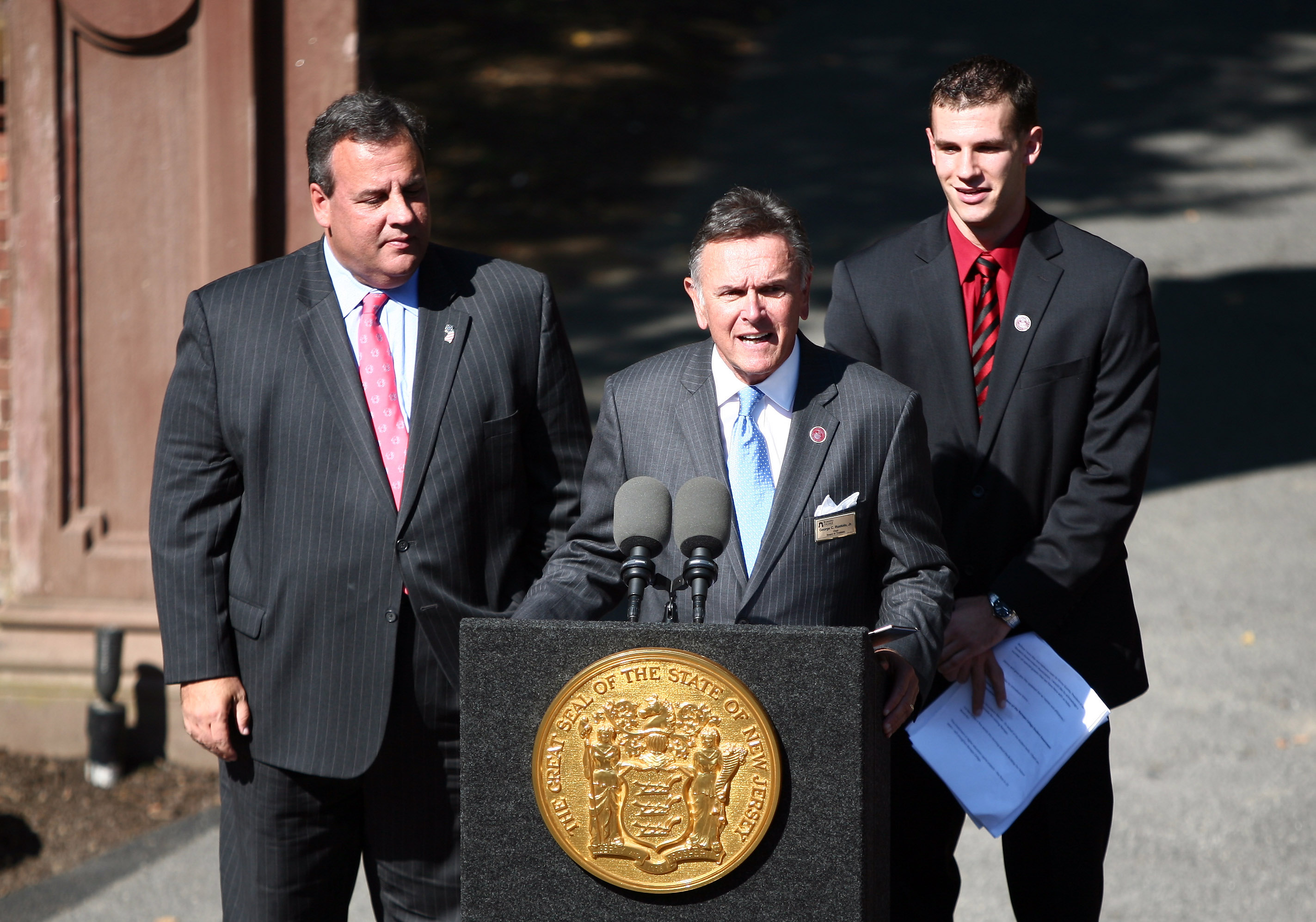 George Ruotolo speaks at a podium at Ramapo College with Governor Chris Christie on the left and student government president Vincent Guinta on the right.