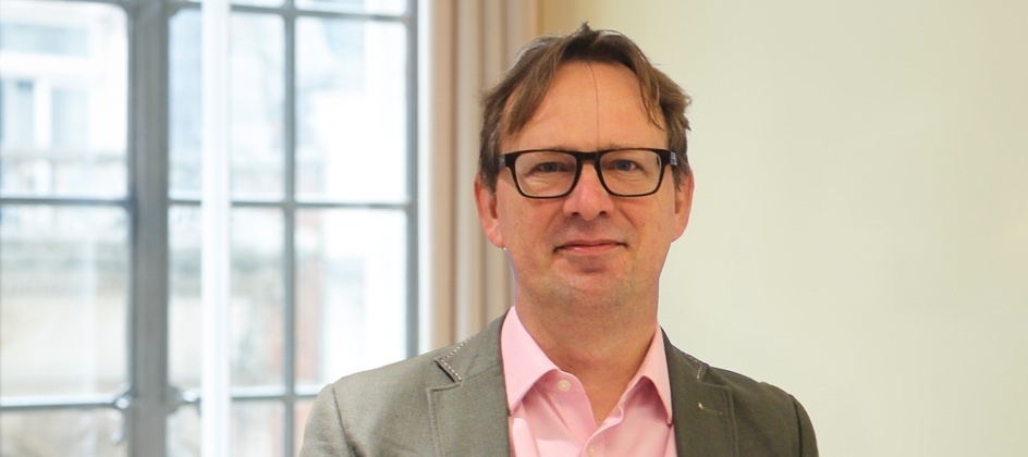 George Coleman wearing glasses, a grey jacket and a pink shirt beside an office window