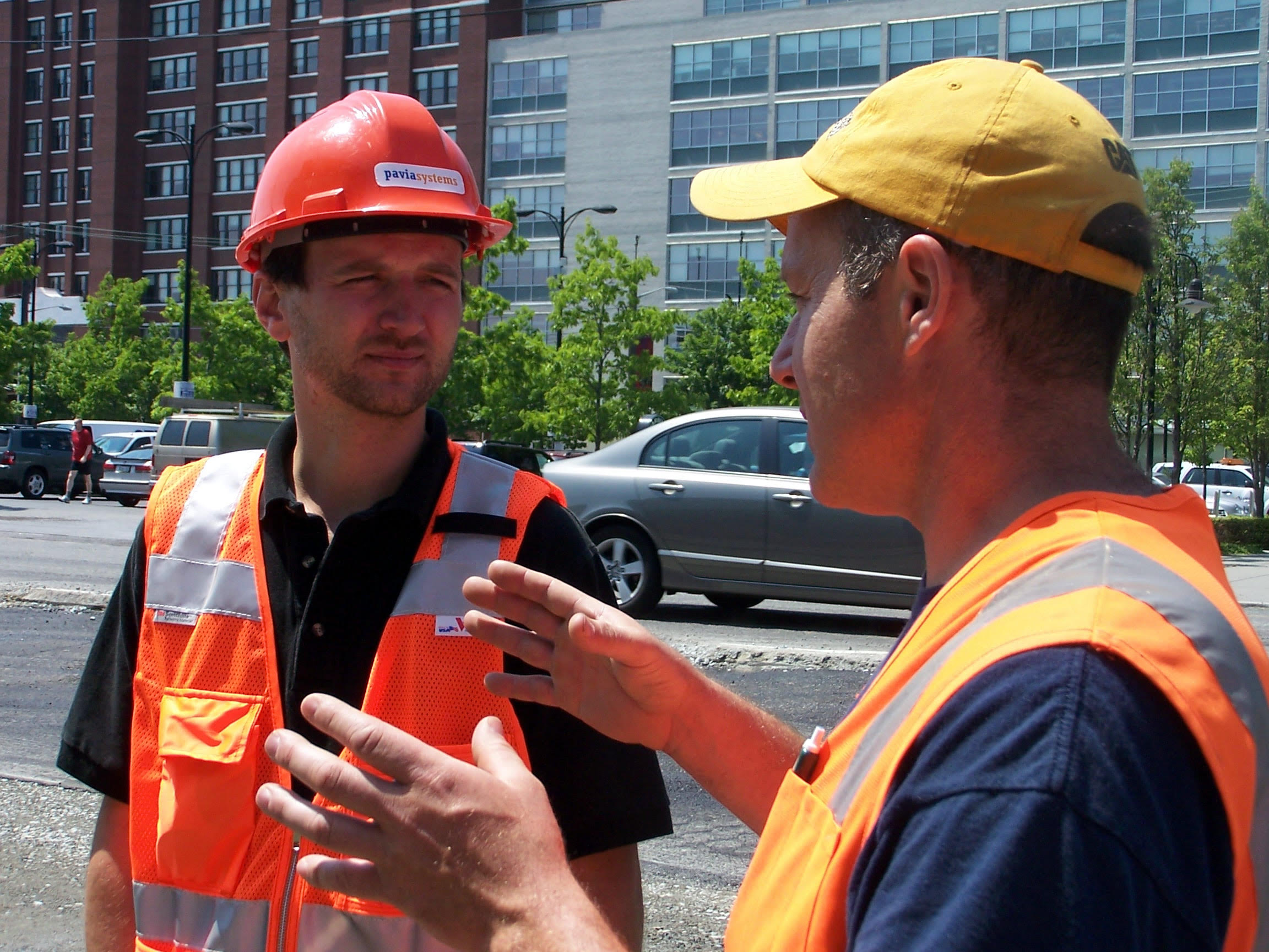 A younger George White in a Pavia Systems hard hat and safety vest listening to a construction worker at a jobsite