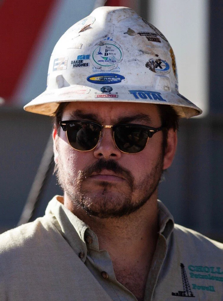 Gideon Powell wearing sunglasses and a sticker-covered hard hat in an oilfield photograph
