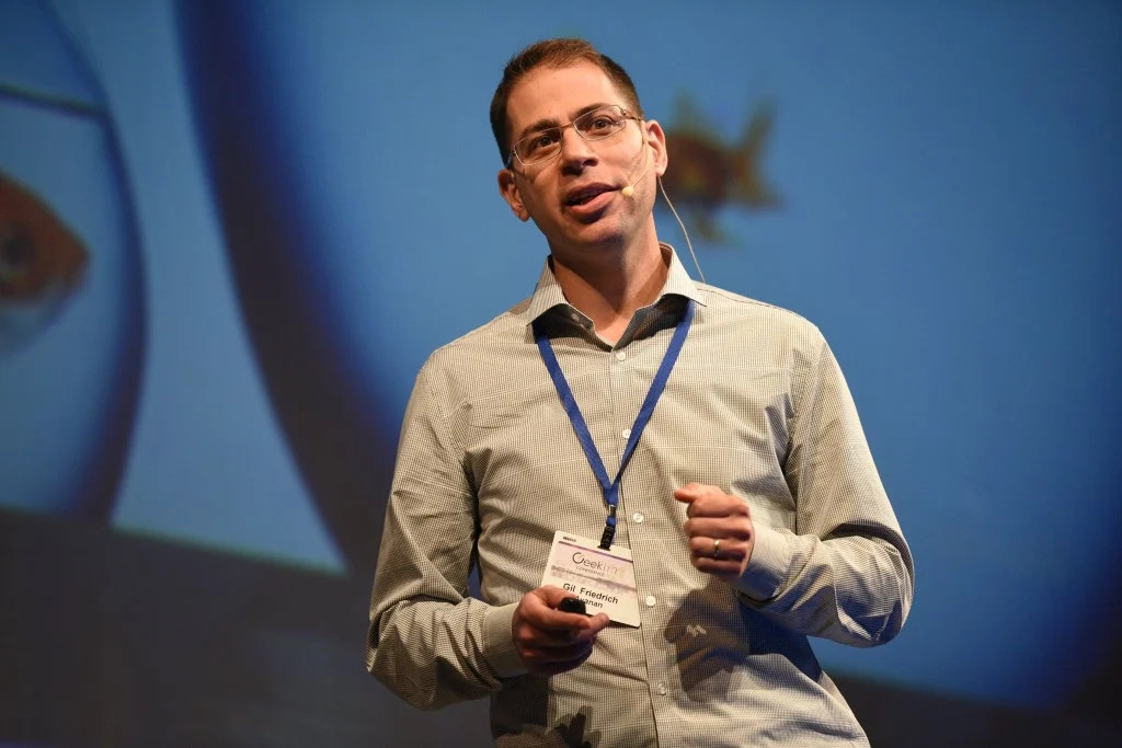Gil Friedrich speaking on stage, wearing a headset microphone and holding a presentation remote