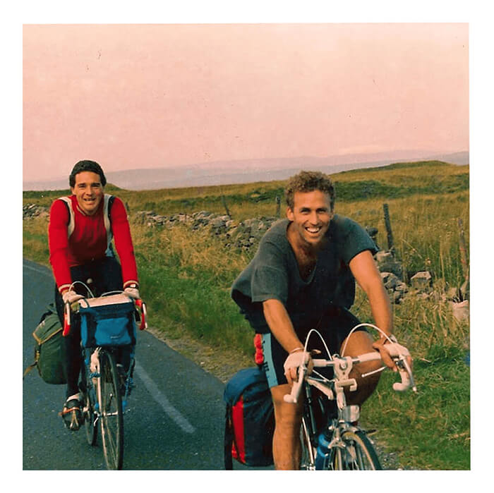 A younger Glen McDermott smiling while riding a loaded touring bicycle beside another cyclist on a country road
