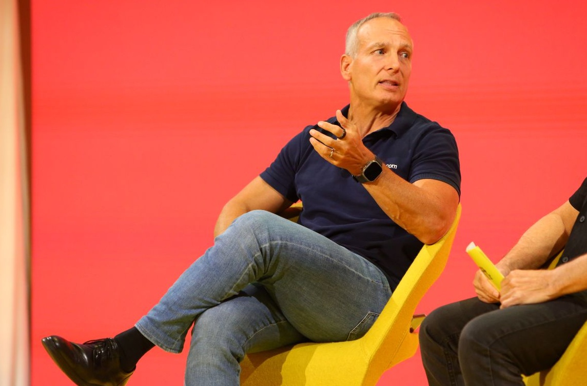 Glenn Fogel speaking from a yellow chair at Skift Global Forum 2025