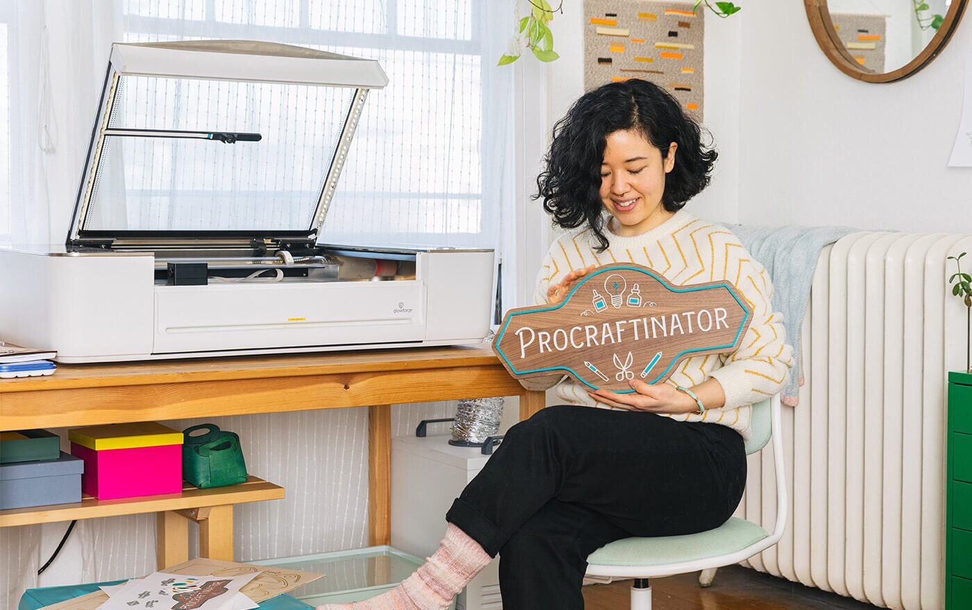 A maker holding a wooden Procraftinator sign beside a Glowforge laser