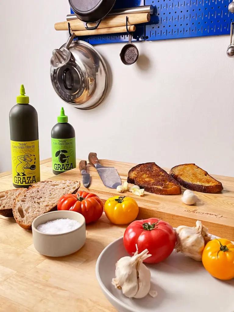 Graza olive oil on a table with tomatoes, bread, garlic and salt