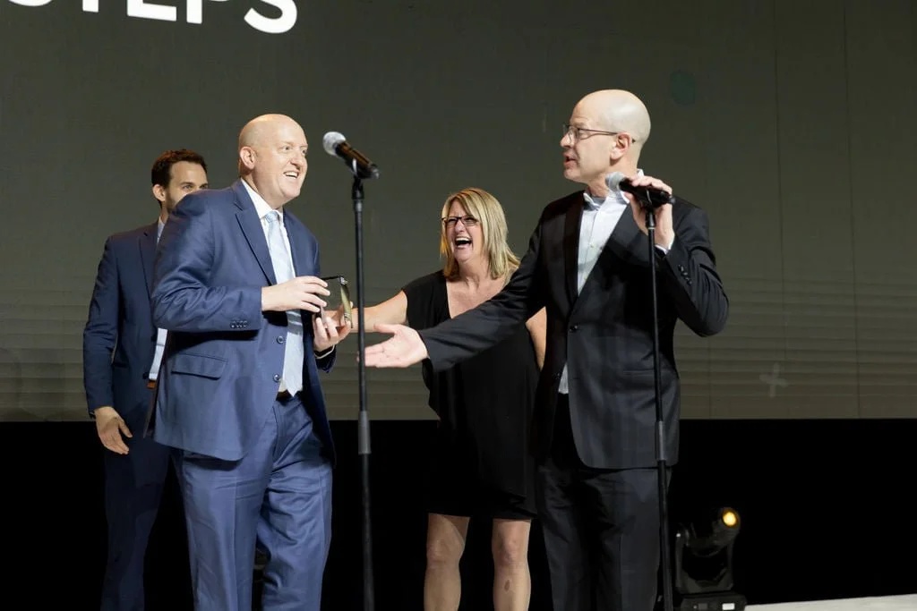 Greg DeVore, in a blue suit, smiling with other people on a stage