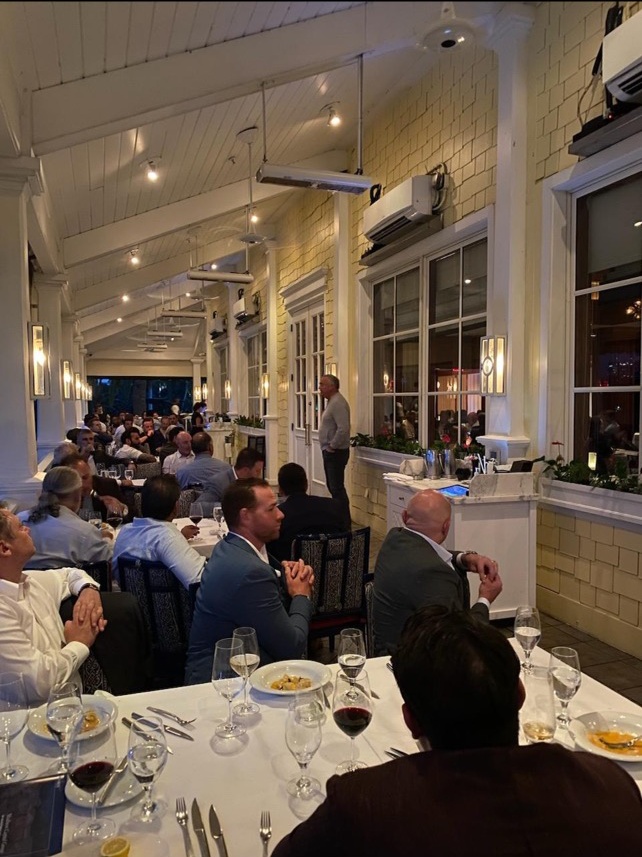 Attendees seated at long dinner tables on a veranda during Coastal’s Palm Beach investor event.