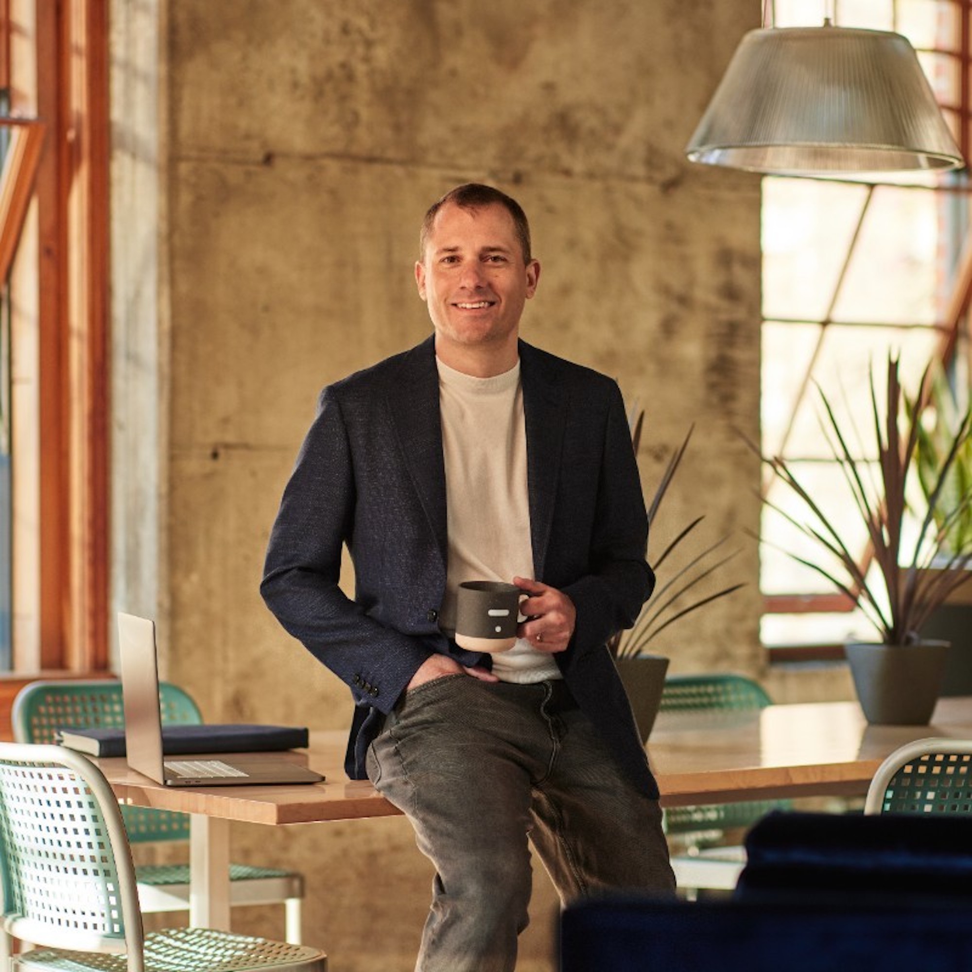 Greg Smith sitting on a table beside a laptop, holding a mug