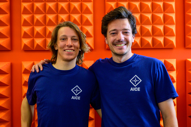 AICE co-founders Rémi Bouteiller and Grégoire Chomette wearing blue AICE shirts against an orange wall