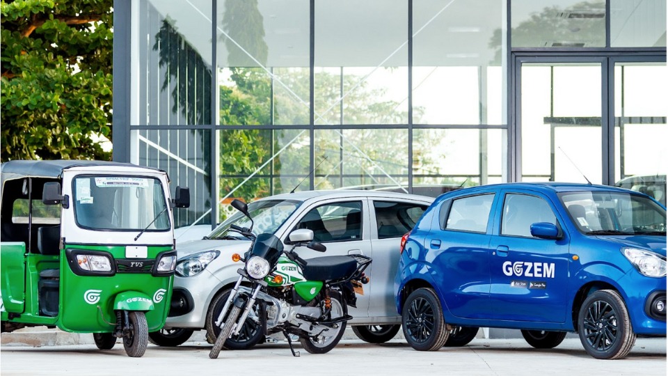 A green Gozem tricycle, motorcycle, silver car and blue Gozem car parked side by side