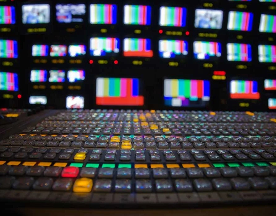 A television production switcher facing a wall of broadcast monitors