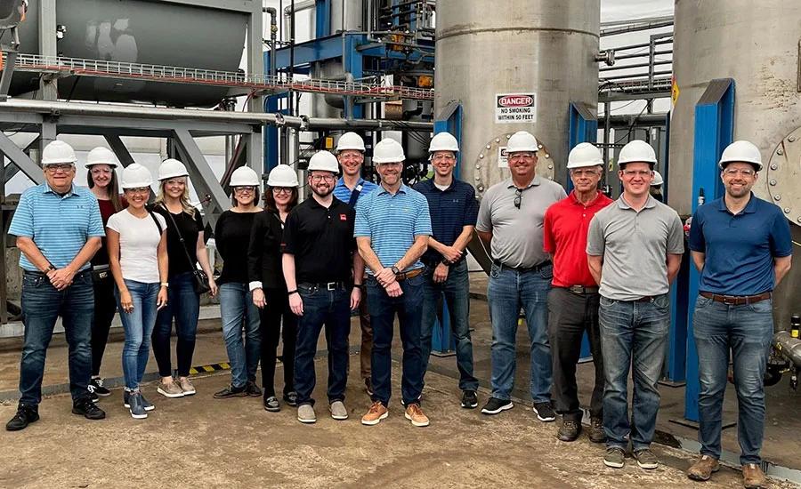 Gunner Smith, at center in a striped blue shirt, with Owens Corning colleagues wearing hard hats inside a manufacturing facility