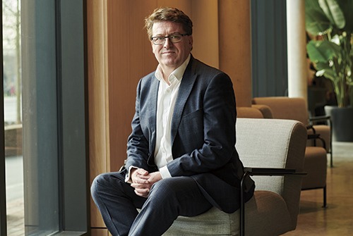 Guy Battle seated beside a window, wearing a dark suit and open-necked white shirt