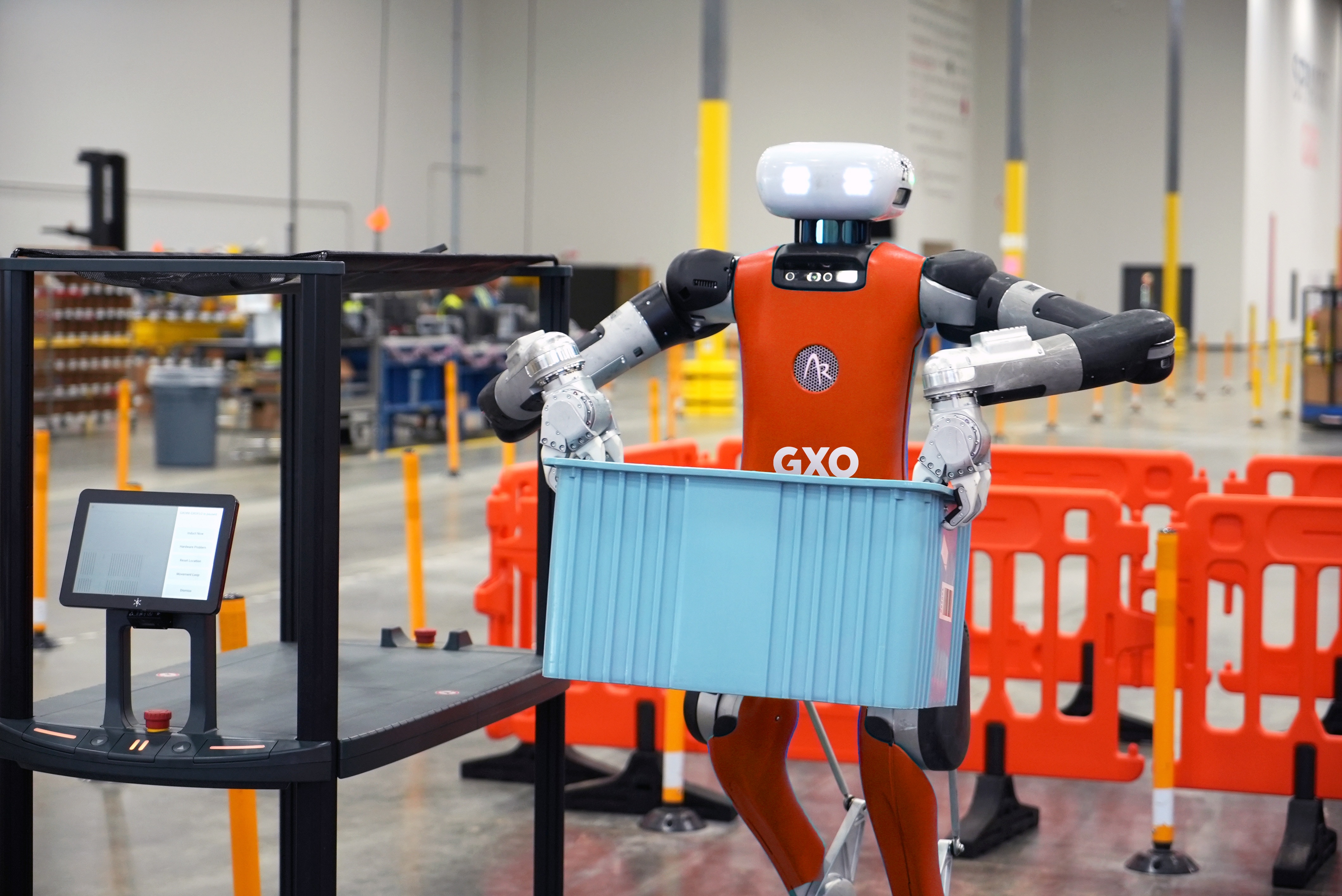 An orange-and-gray Digit robot grips a blue tote beside a warehouse conveyor