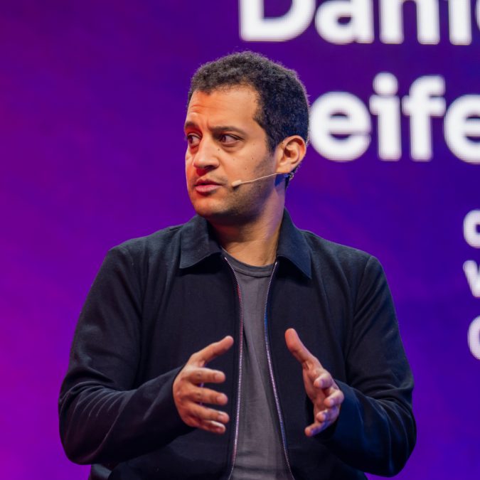 Hany Rashwan speaking onstage with a headset microphone against a purple backdrop