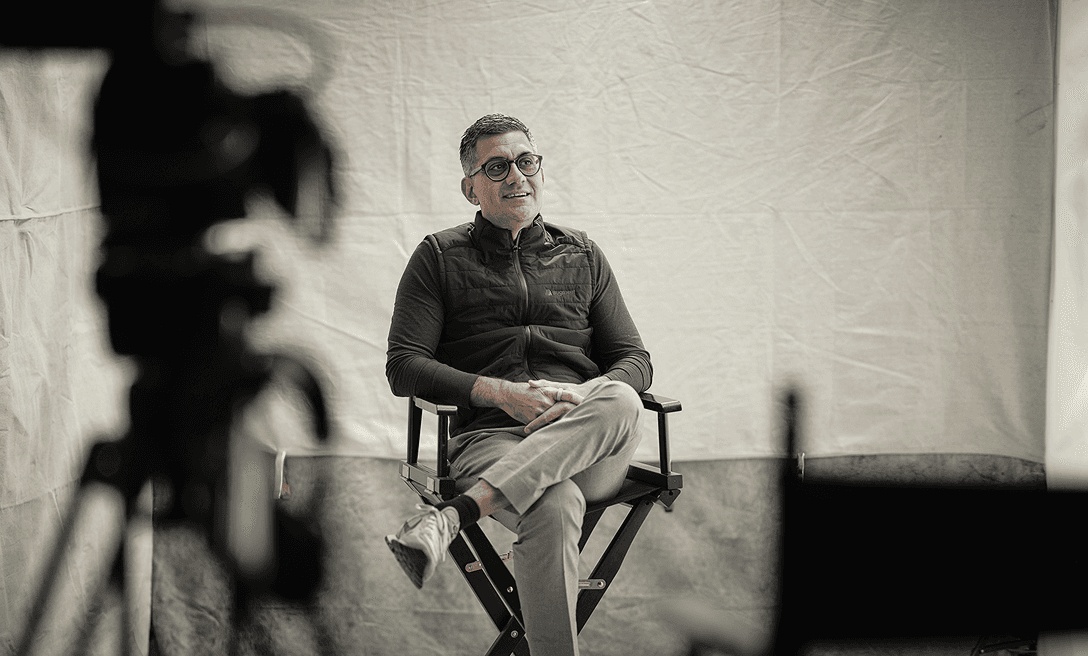 Harish Abbott seated in a director's chair during filming, with camera equipment in the foreground