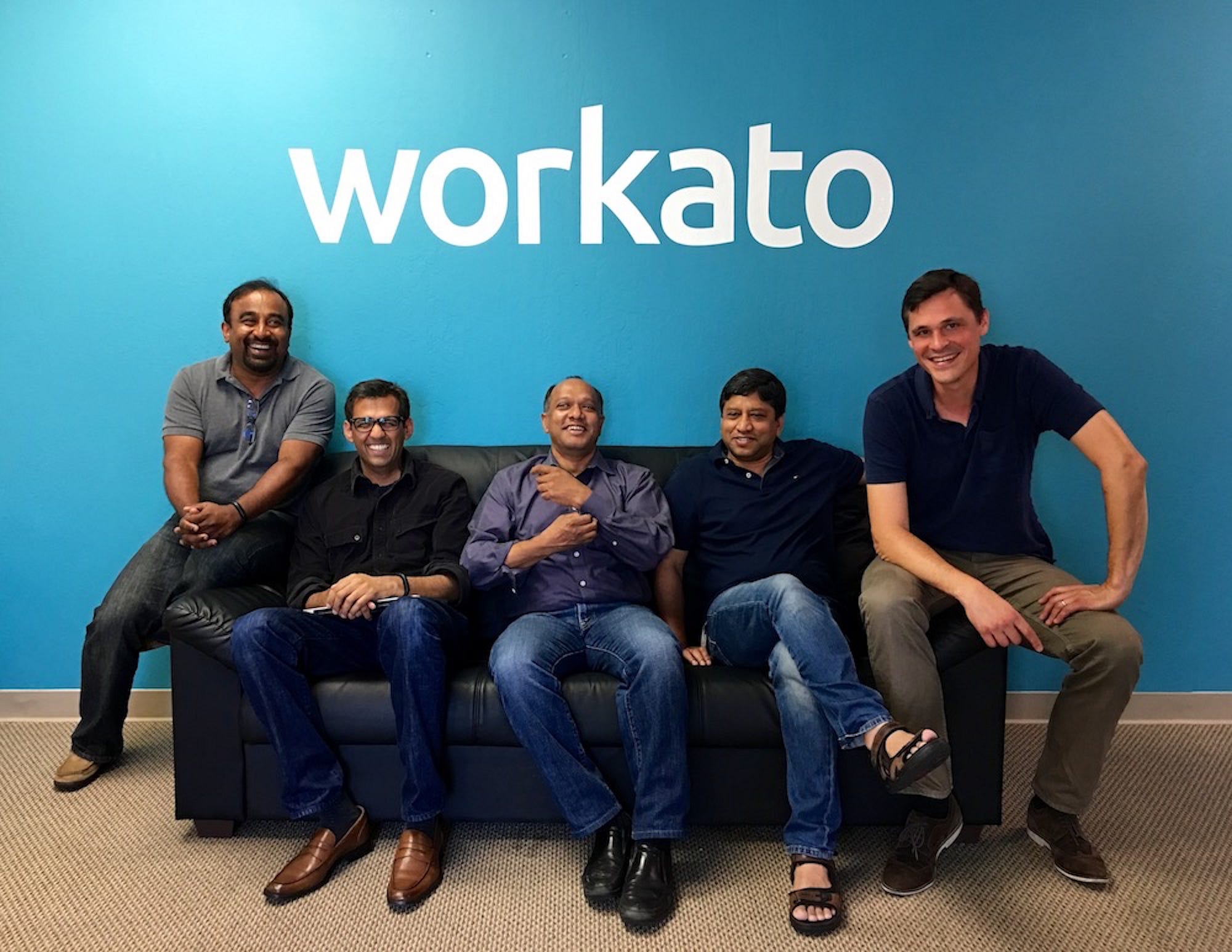 Five members of Workato's early team seated together beneath a large Workato sign