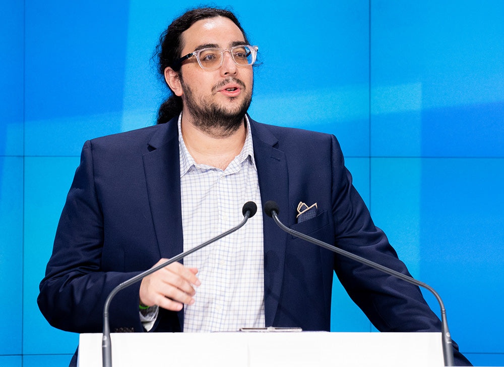 Harrison Gross wearing clear-framed glasses and a navy jacket while speaking at a lectern