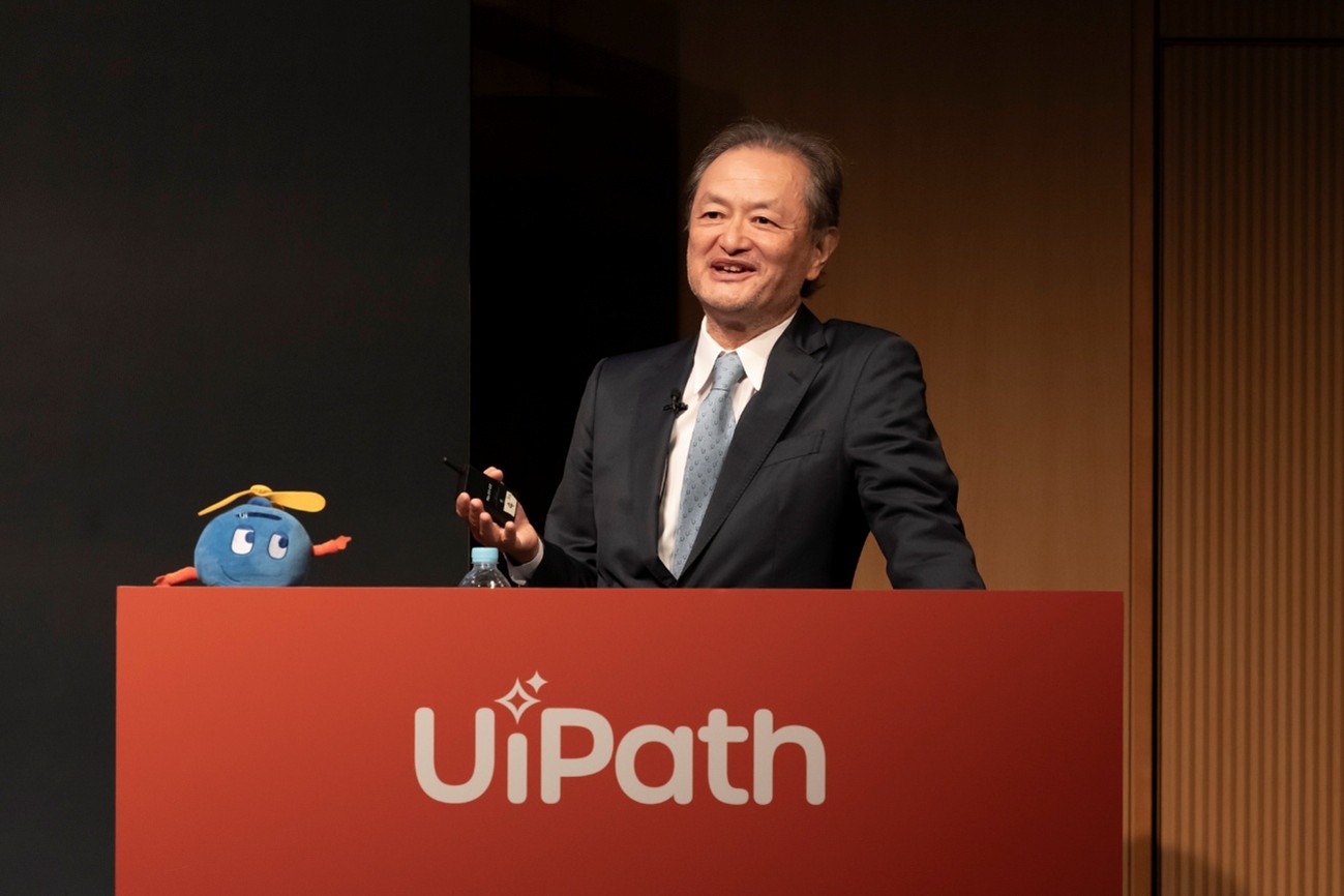 Koichi Hasegawa speaking behind a UiPath lectern at the 2025 partner summit, beside a small blue mascot