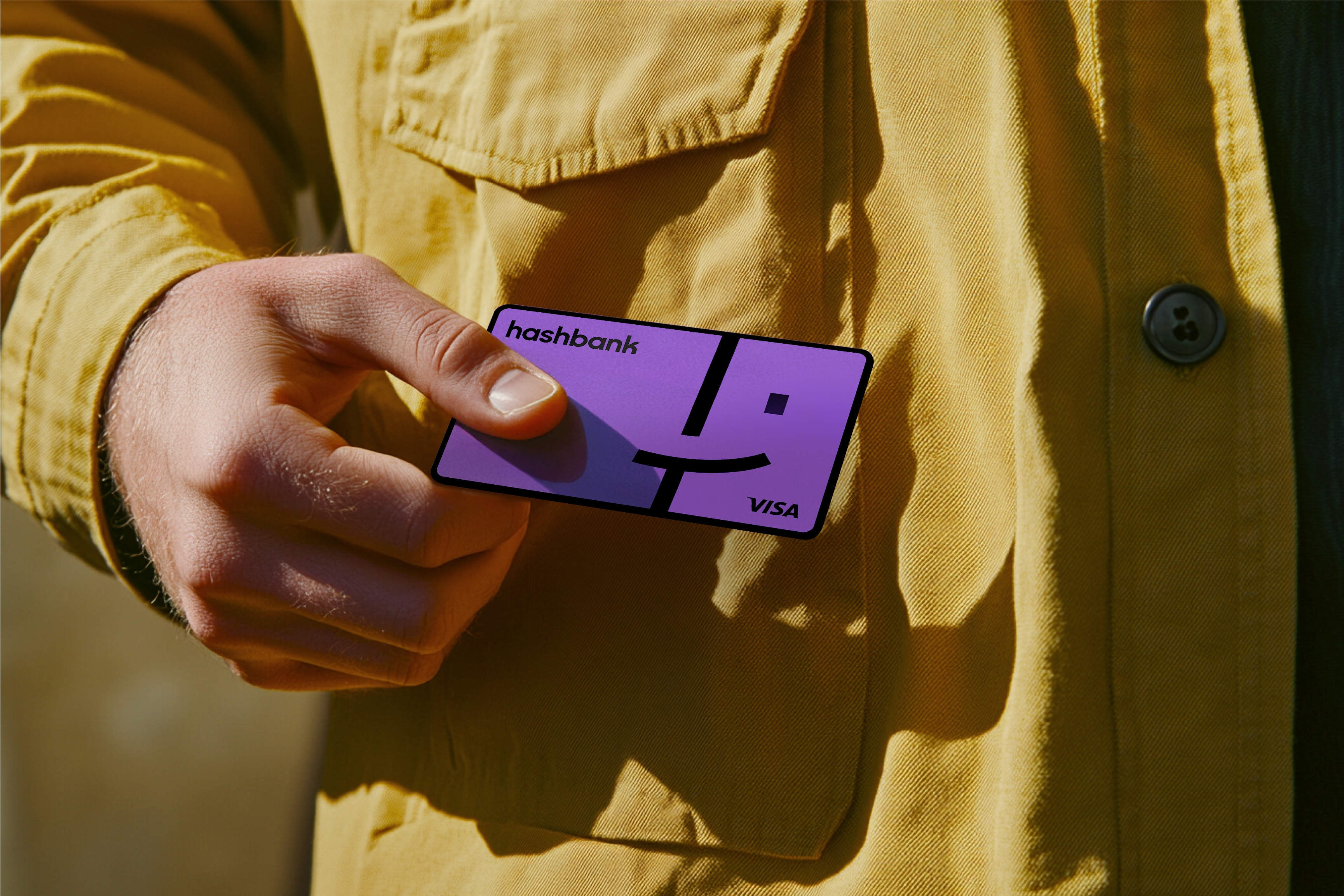 A hand holding Hashbank’s purple Visa card against a mustard-yellow jacket