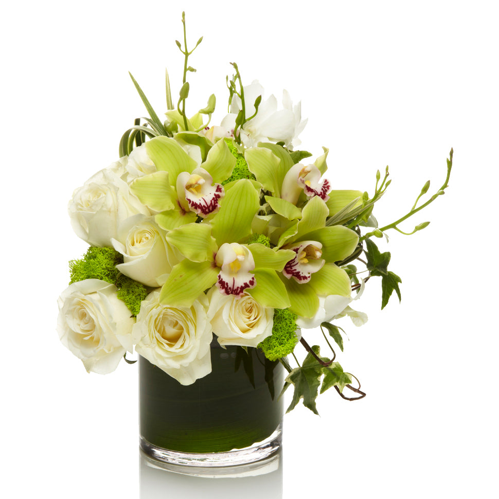 H.Bloom arrangement of white roses, green orchids and foliage in a glass vase