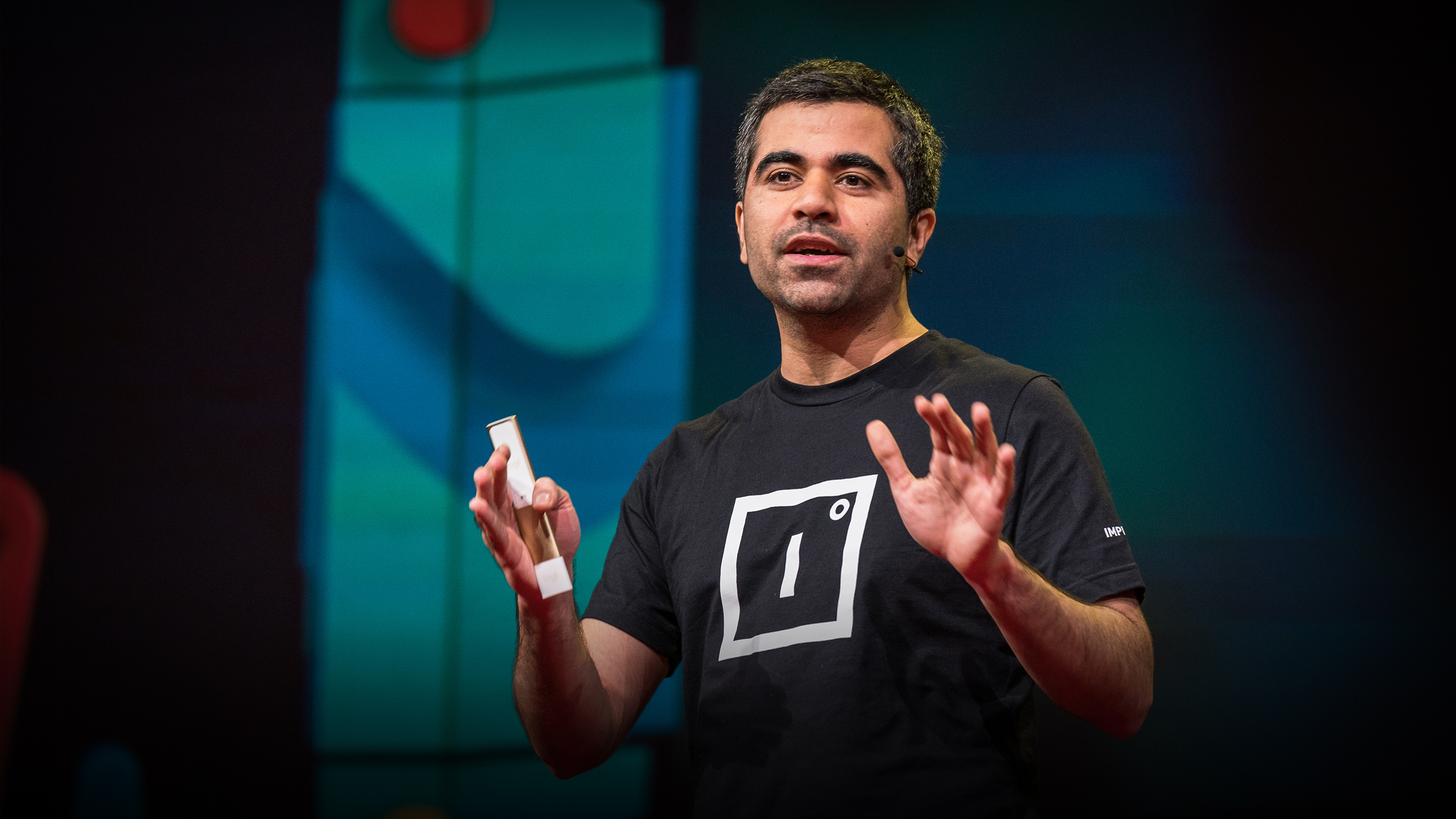 Herman Narula speaking on the TED stage in 2019, wearing an Improbable T-shirt and gesturing with both hands
