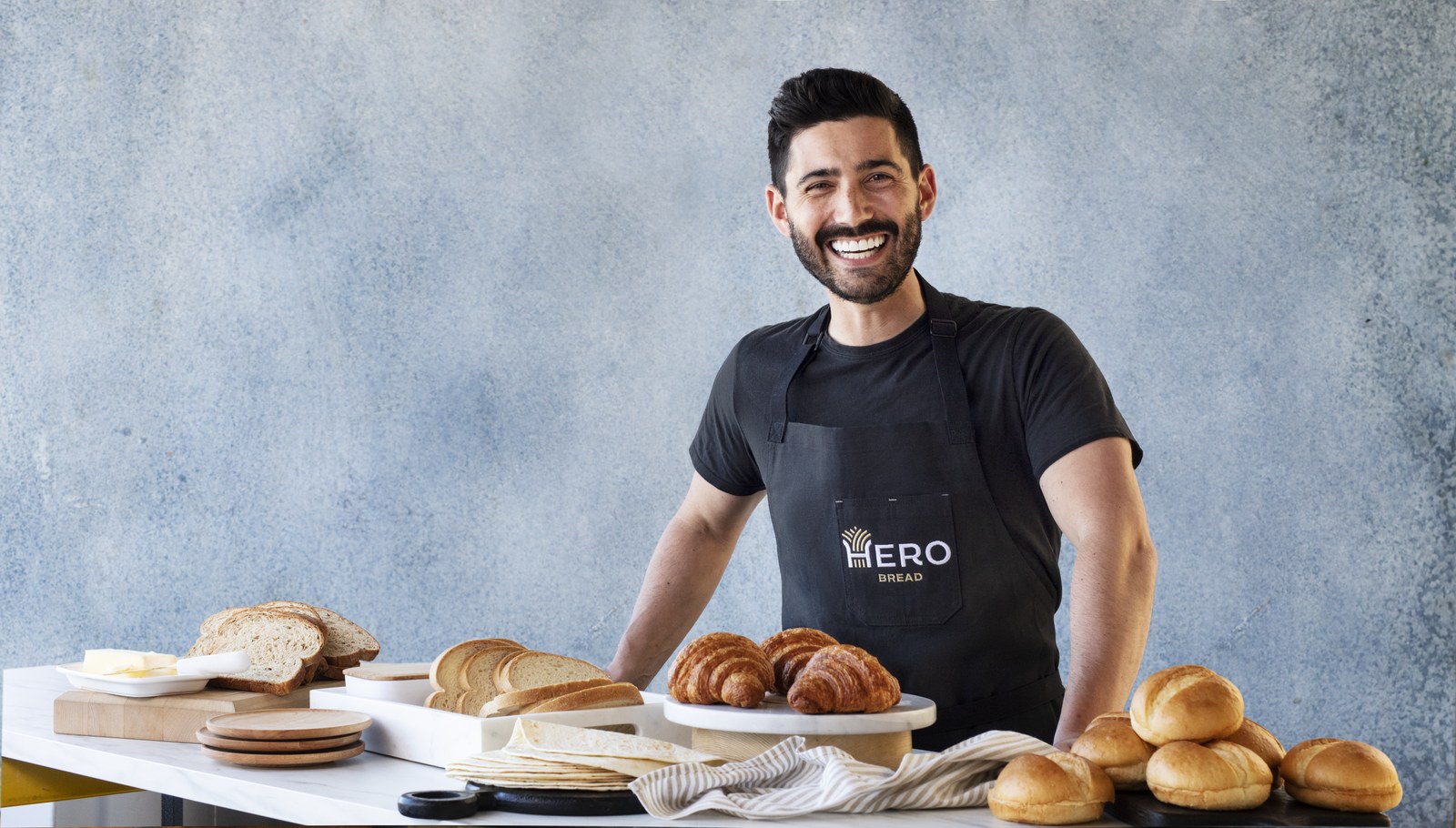 Hero Bread founder Cole Glass standing behind breads, tortillas, rolls and croissants