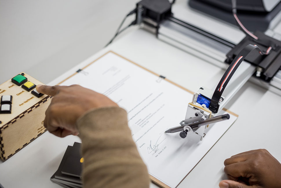 A signature-writing robot holds a pen over pension paperwork as an operator presses a button.
