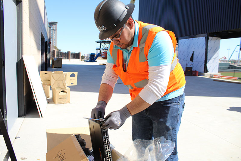 A worker in a hard hat and orange safety vest handles network equipment during Riverfront Stadium construction