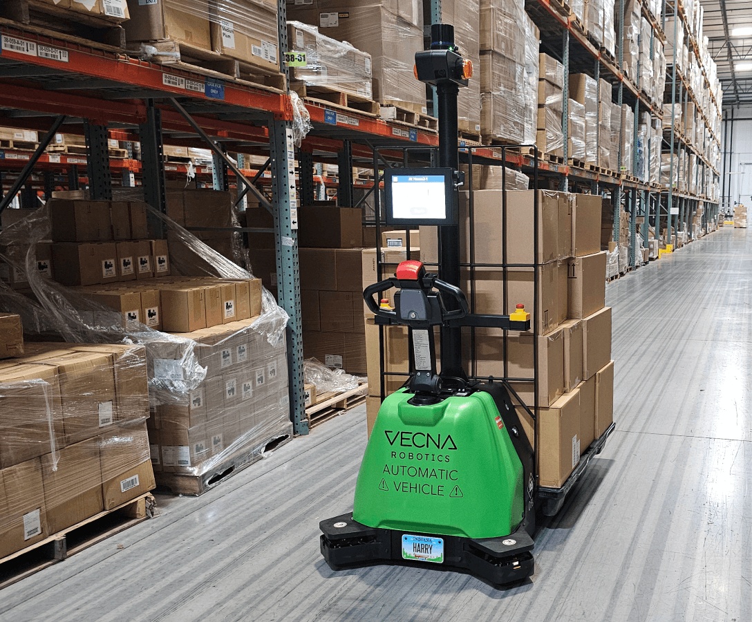 A green Vecna Robotics automated pallet jack carrying boxed goods down a warehouse aisle
