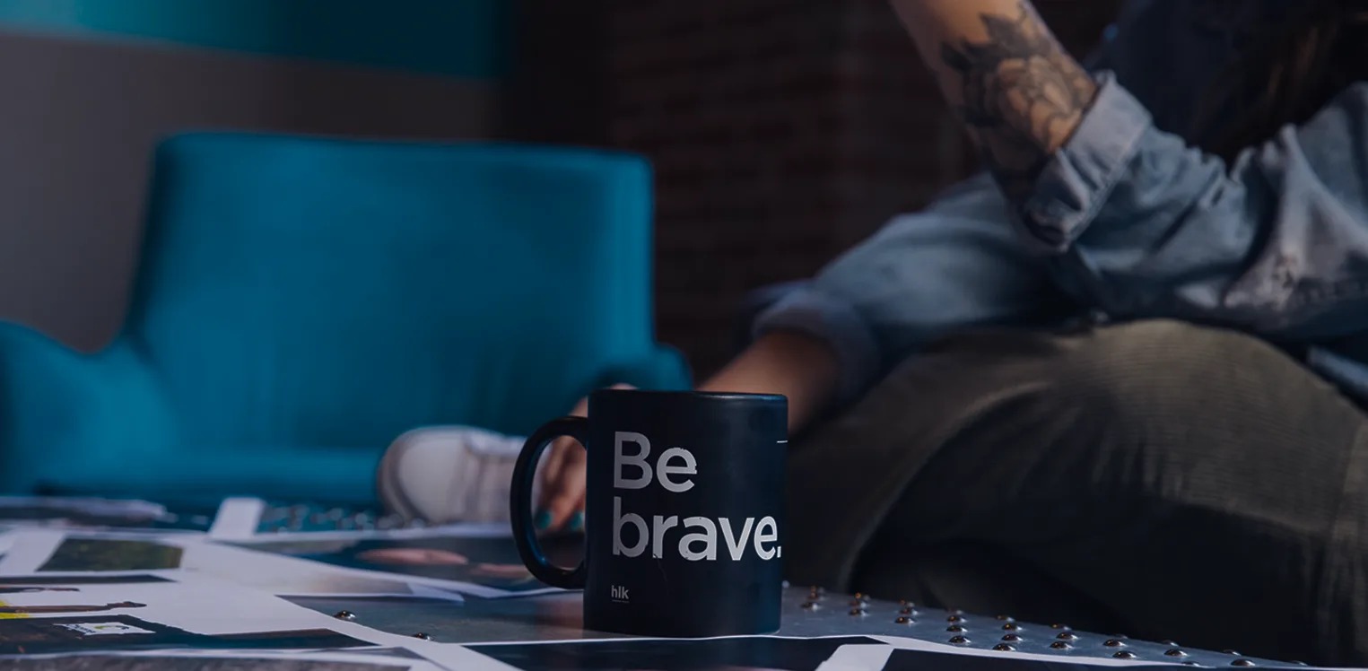A dark HLK mug printed with Be brave sits among creative materials in the agency office