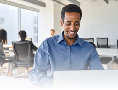 A smiling office worker seated at a laptop in a bright shared workspace.