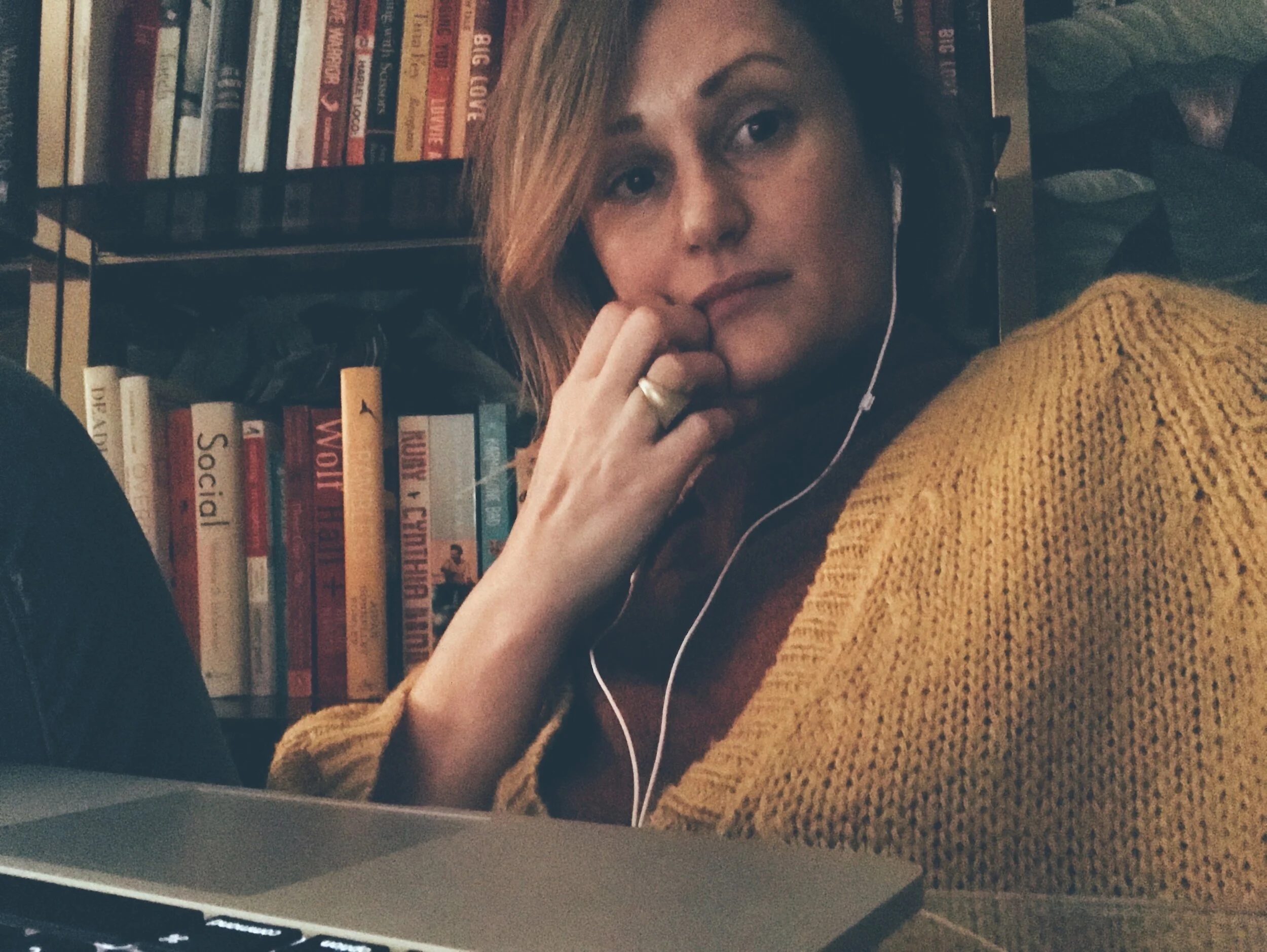 Holly Whitaker in a yellow cardigan, wearing earphones beside bookshelves with a laptop in front of her
