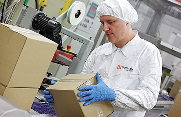 A worker in a Holman-branded uniform and gloves handles a cardboard box beside production equipment