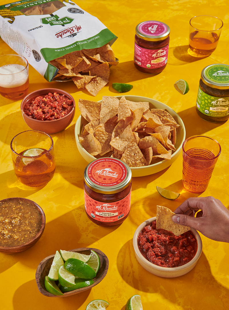 Mi Rancho salsa jars, tortilla chips, and drinks arranged on a yellow table