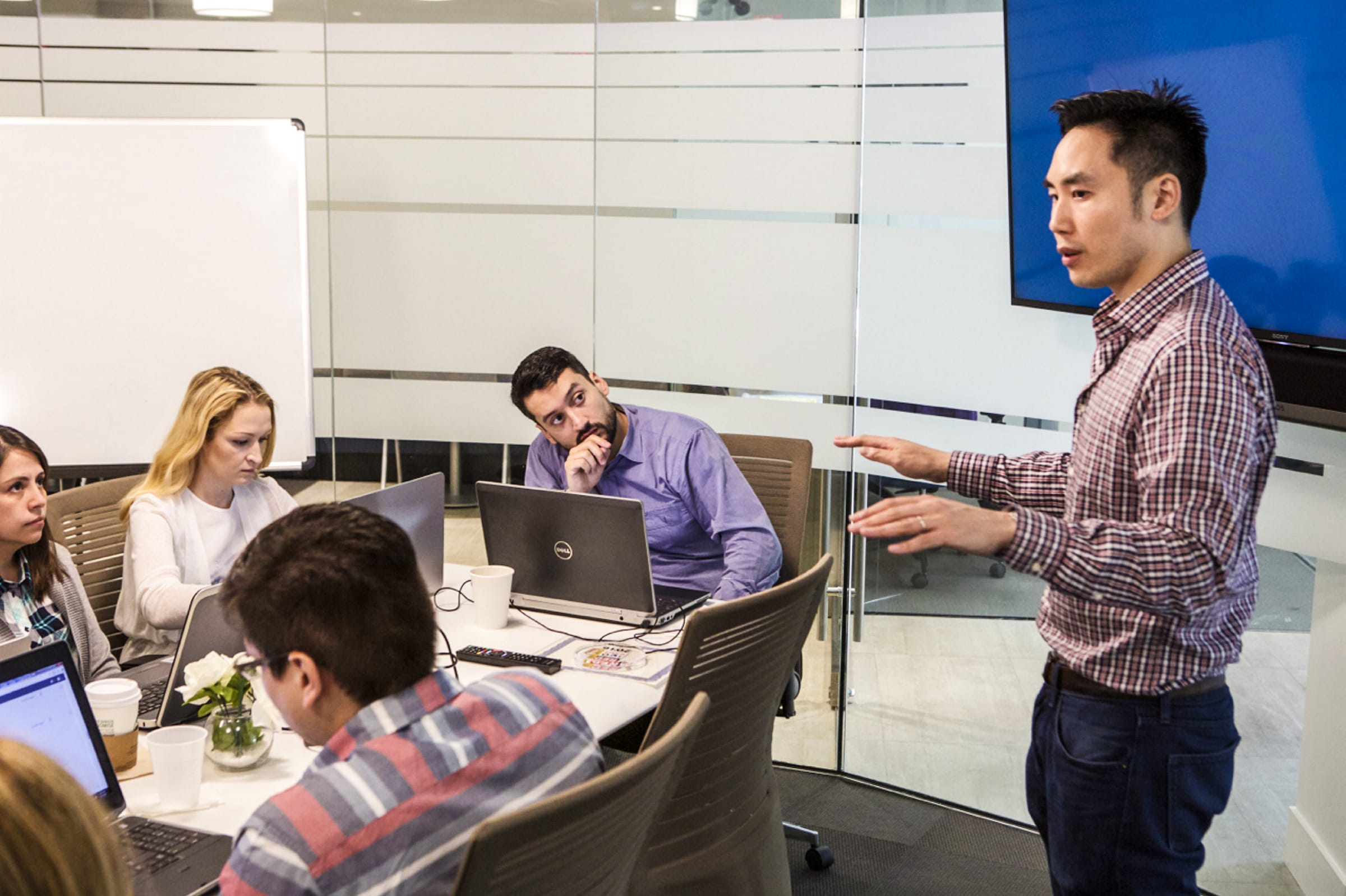 Howard Yeh standing beside a conference table and speaking with colleagues working on laptops