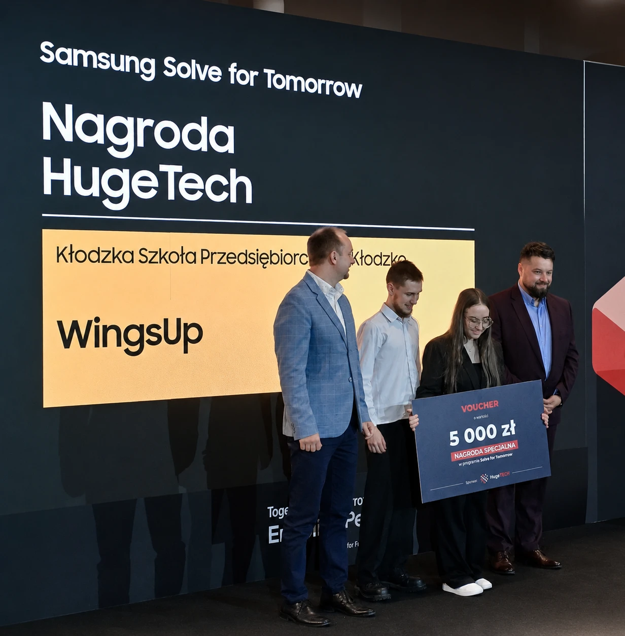 Recipients holding a 5,000 PLN voucher beneath a HugeTech award sign at Samsung Solve for Tomorrow