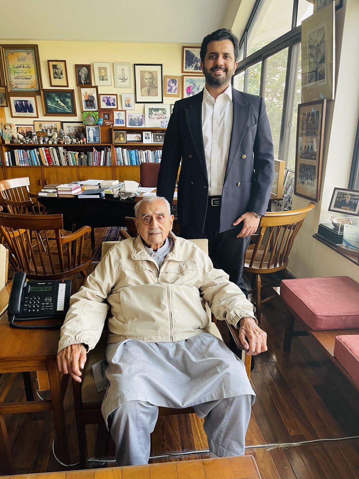 Hussain Nasir standing beside a seated Syed Babar Ali in a room lined with books and framed photographs