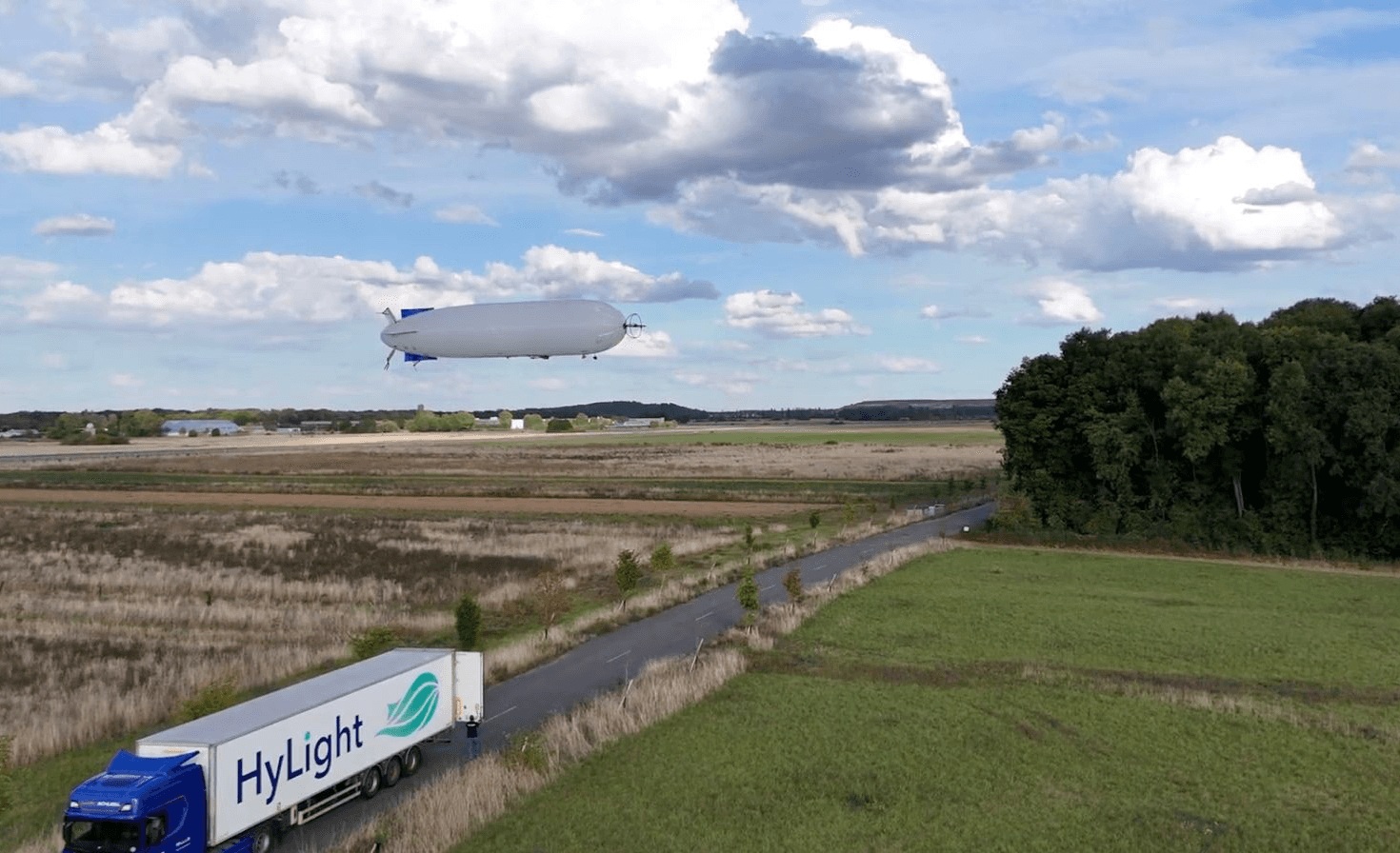 A HyLight airship flies above a field beside its branded support truck