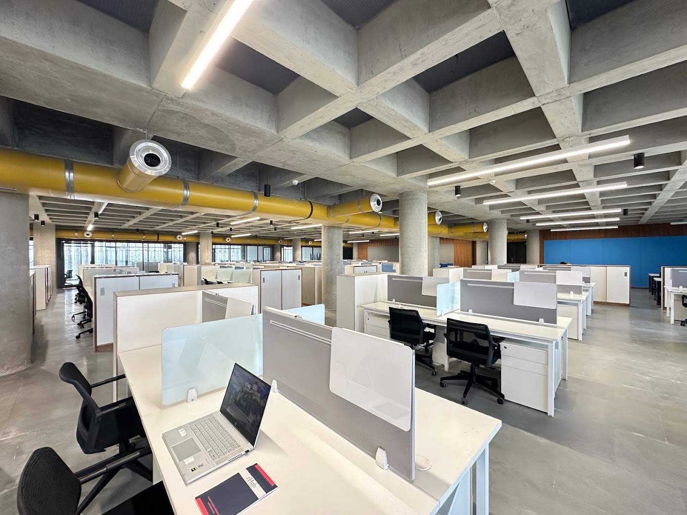 Co-working desks beneath exposed concrete beams and yellow ventilation ducts at i-Hub Gujarat
