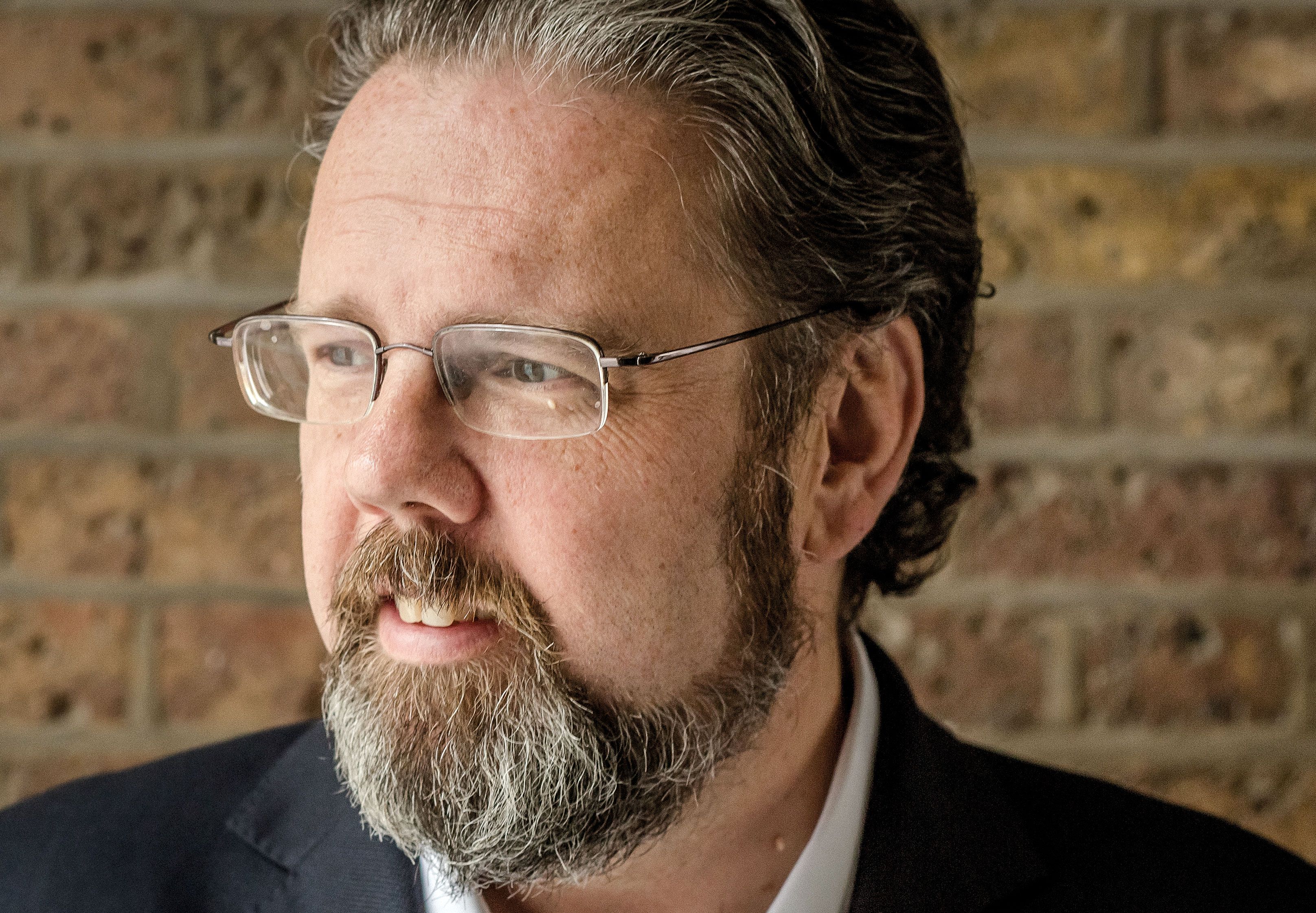 Ian Taylor wearing glasses and a dark jacket, photographed beside a brick wall