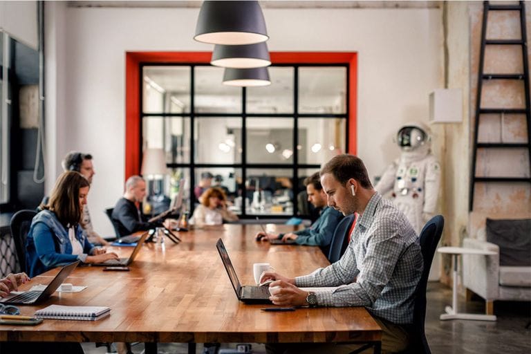 People working at laptops around a shared wooden table in ICT Hub’s coworking space
