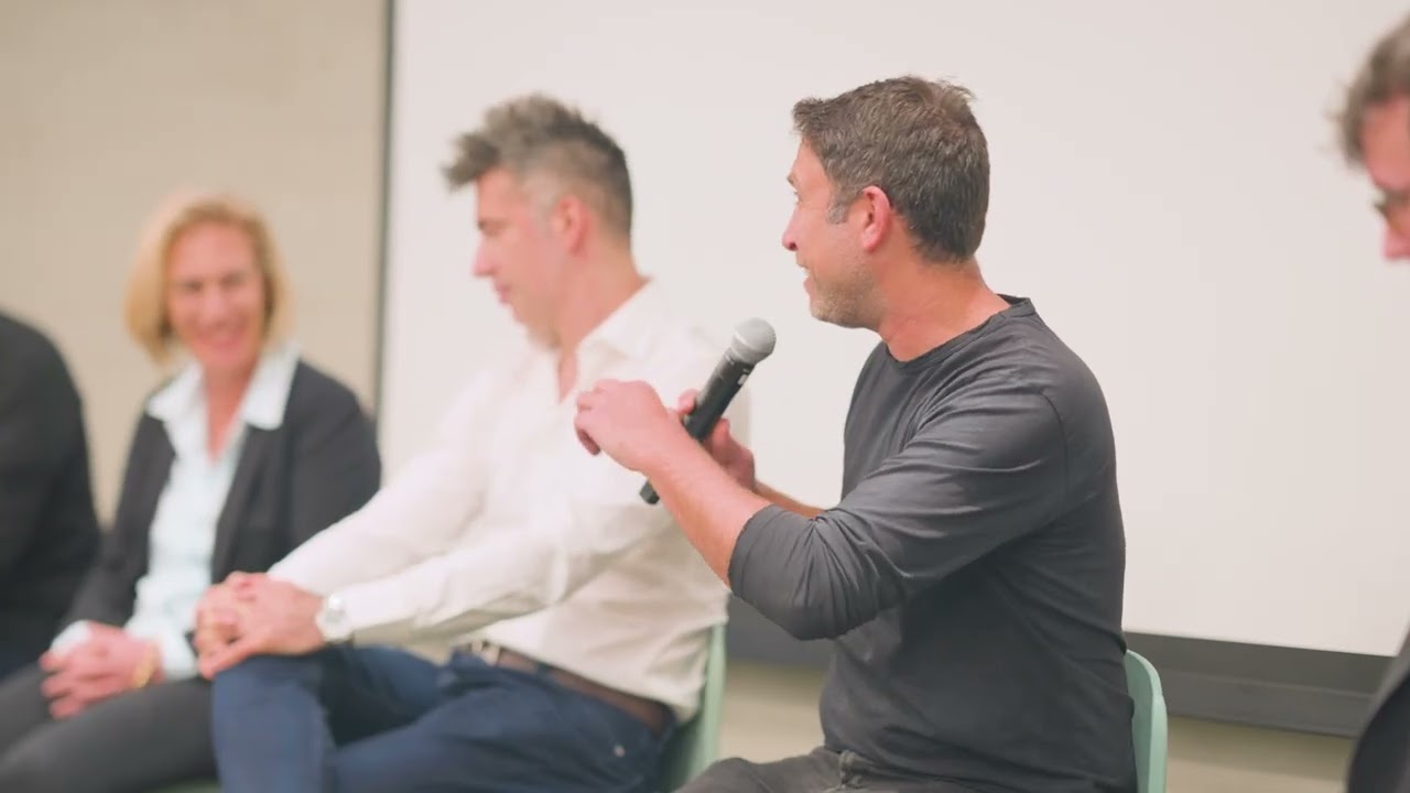 Idan Plotnik speaking into a microphone during an executive panel, with other panelists seated beside him.
