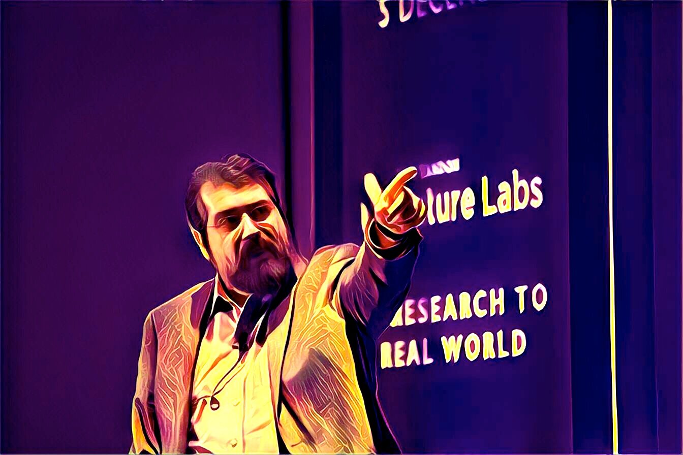 Igor Jablokov gesturing onstage beside a Research to Real World sign, in an image with purple and gold tones