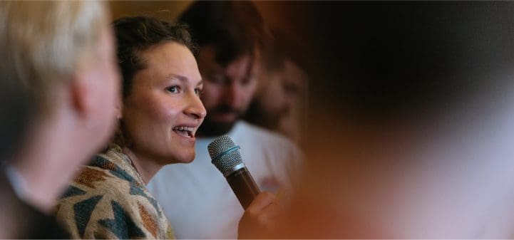 A participant speaks into a microphone among a group at an Impact Hub event.