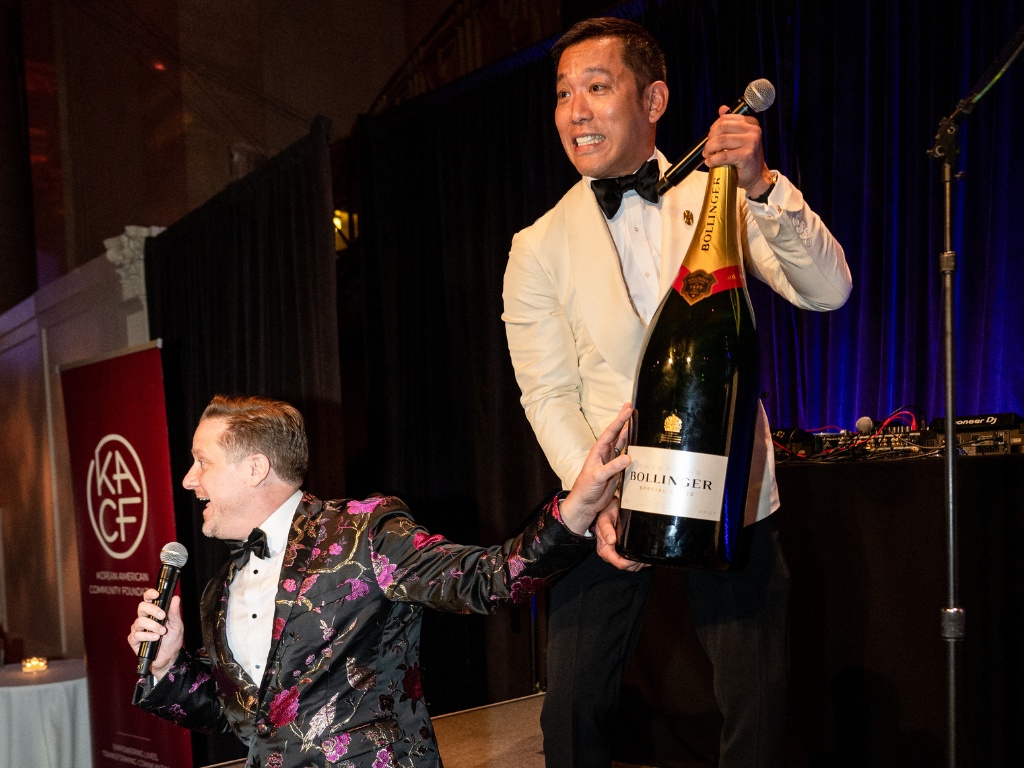 In Seon Hwang in a white dinner jacket holds an oversized champagne bottle and a microphone onstage at KACF’s 2024 gala.