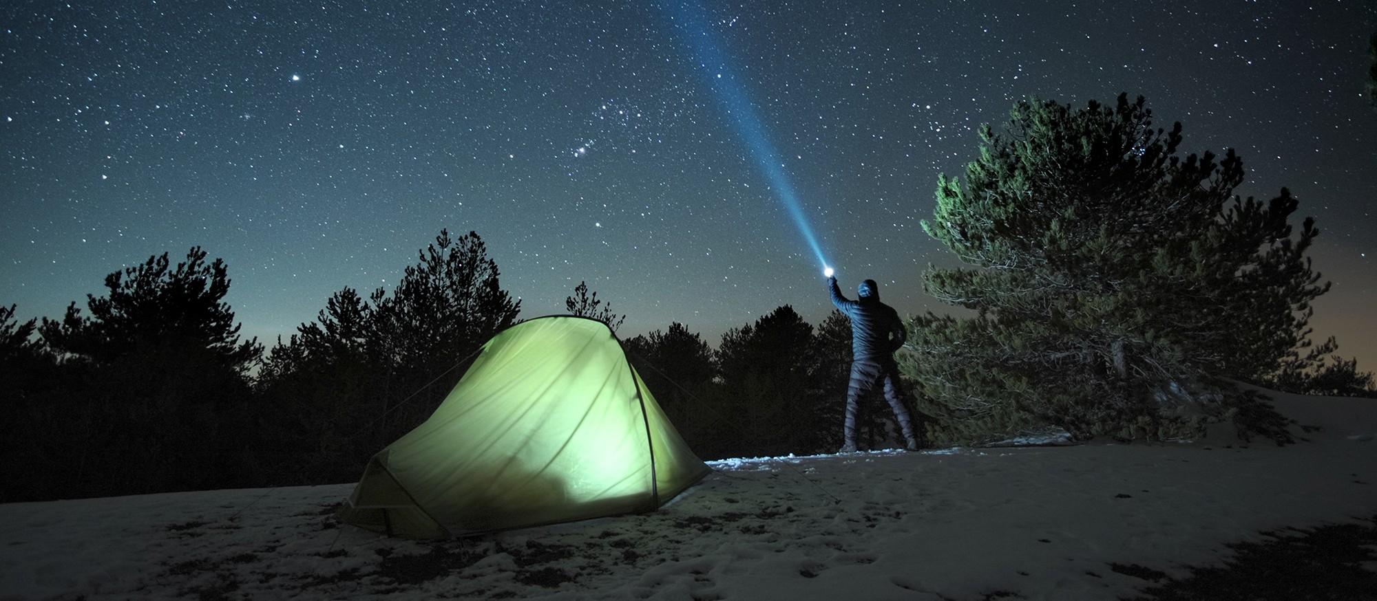 A camper points an Infinity X1 flashlight beam into a starry night sky beside a glowing tent